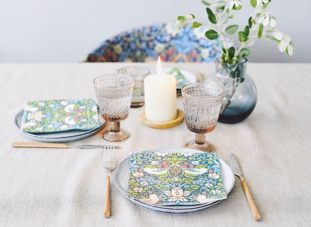 William Morris Paper Napkins with his Strawberry Thief design.   Shown in a dinner table setting with the napkins laid out on the plates. Candles and flowers also adorn the table