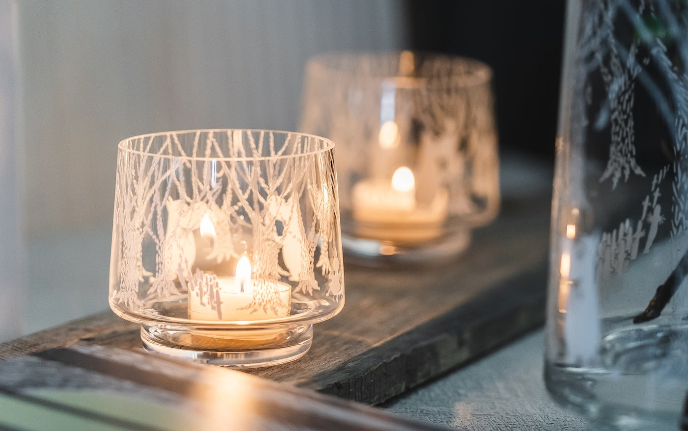2 lit Moomin In The Woods Glass tea light holders on some wooden decking. A beautiful soft warm glow emanating from both.