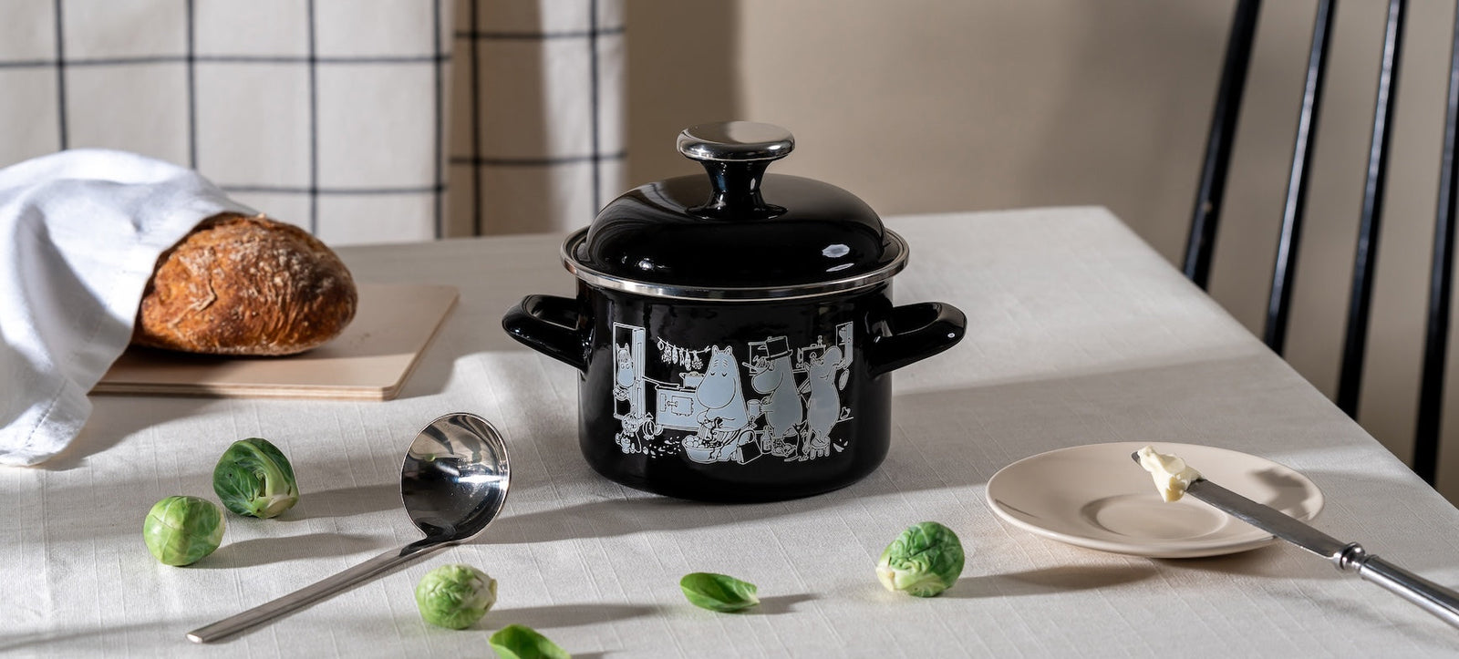 Moomin In The Kitchen enamel cookware by Muurla Design. A black enamel pot with lid shown on a dining table next to bread and sprouts!
