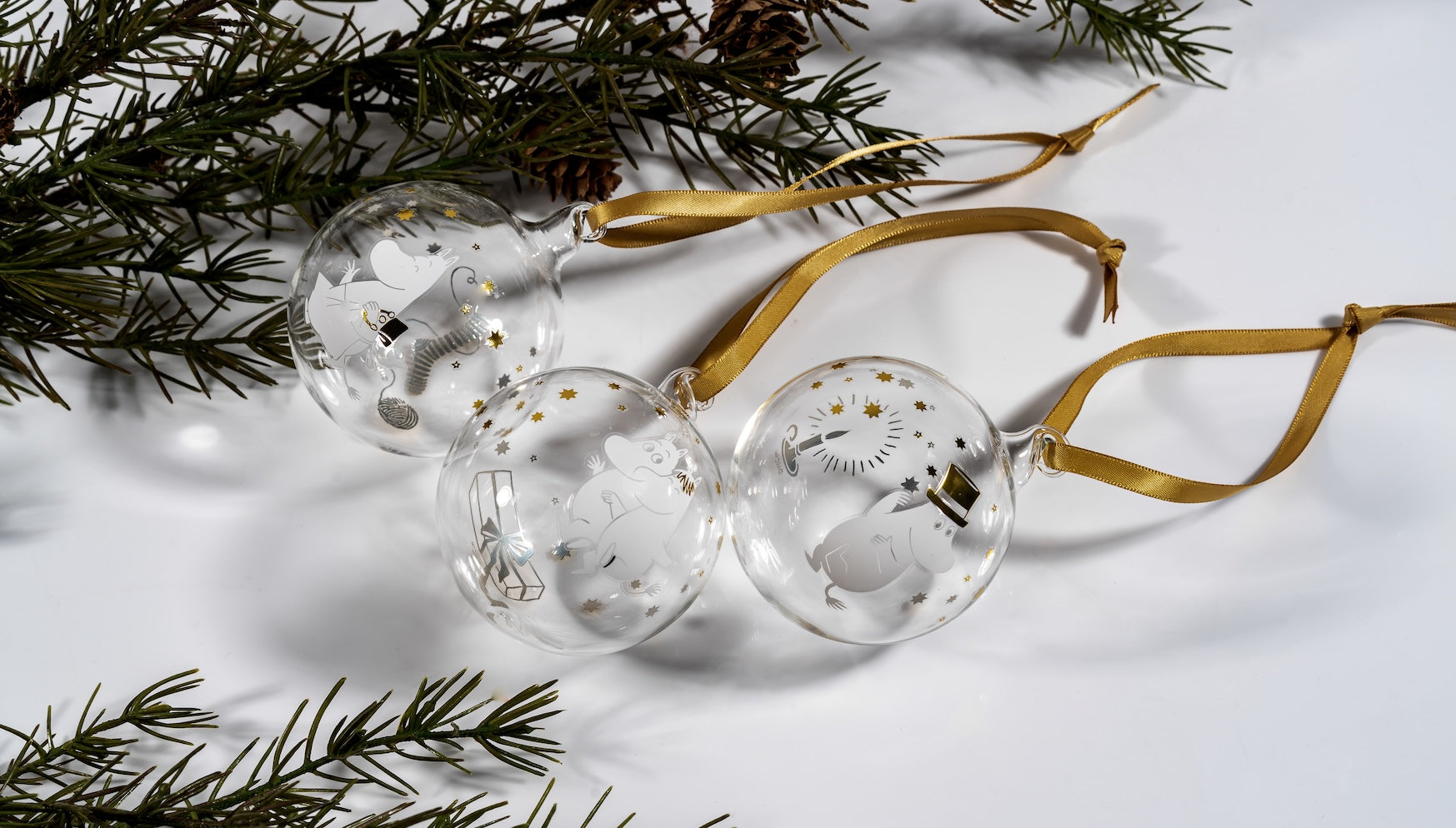 3 Moomin Christmas Glass Baubles with gold ribbons, laying on their side next to Nordic Spruce Fir branches 