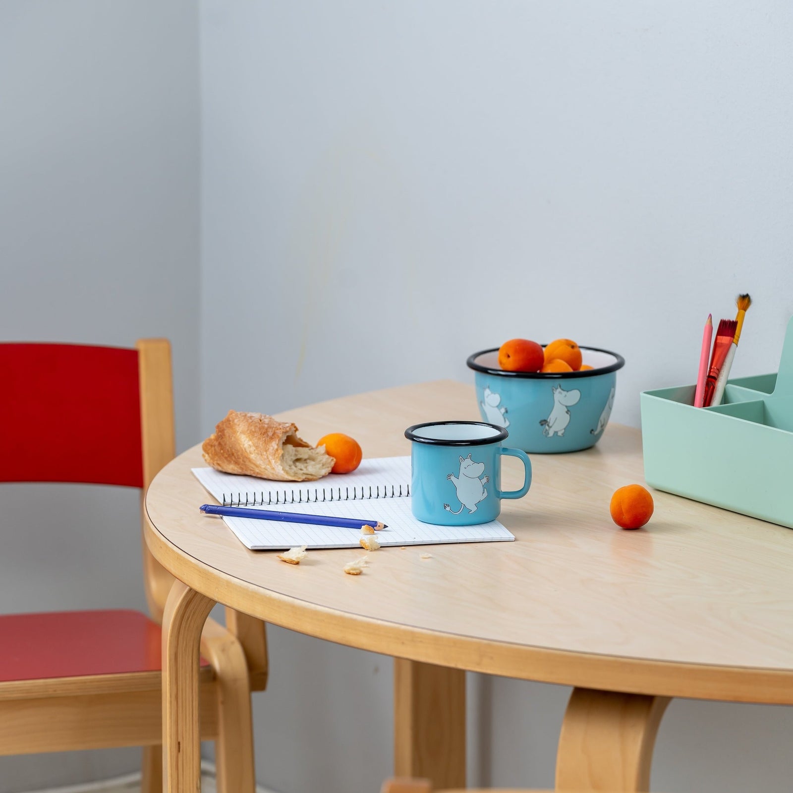 Moomin Enamel Retro Mug in Blue.  Moomintroll Design. Shown on a table with a Moomin enamel bowl and writing pad and pencil