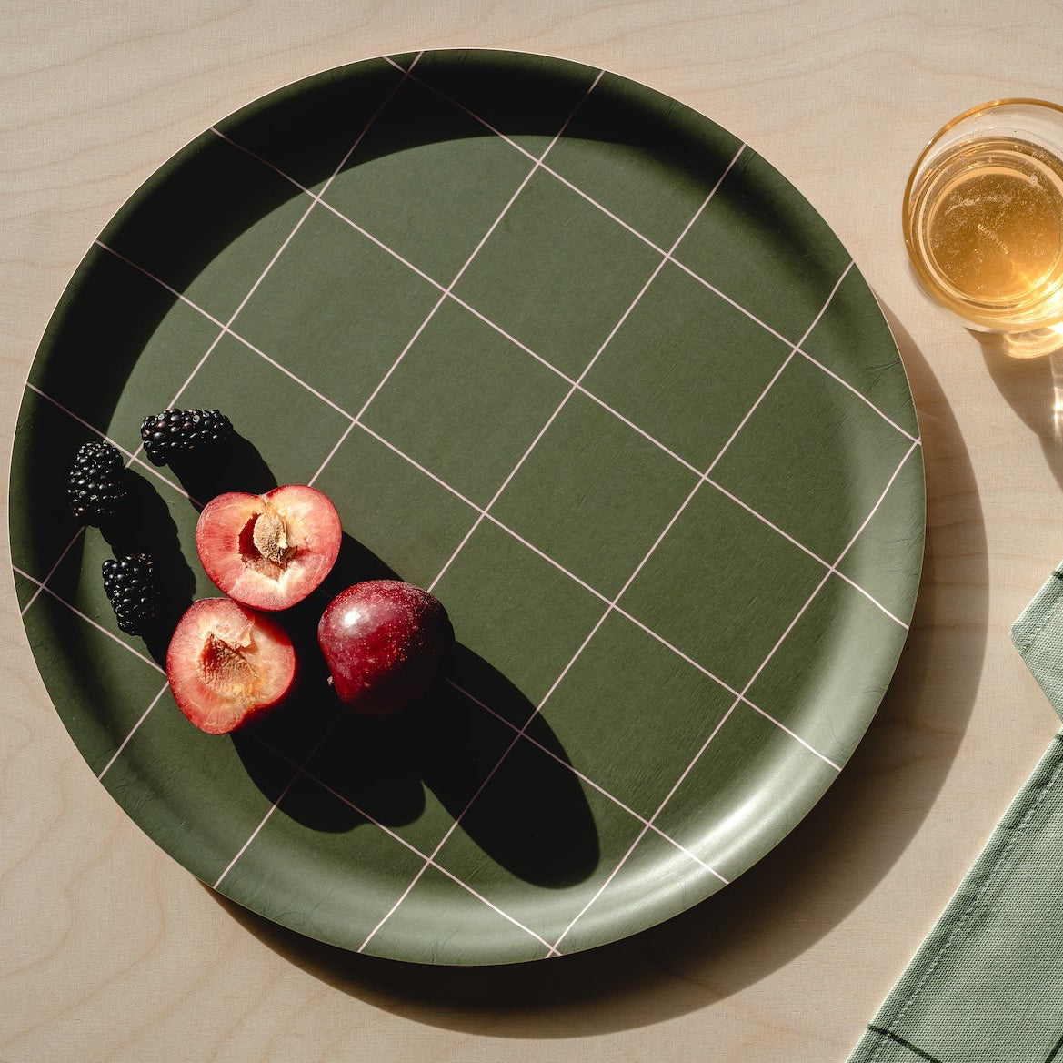 A dark green birch tray with a white grid pattern, styled on a light wood table with fresh fruit, a glass of white wine, and a matching green checkered cloth napkin.