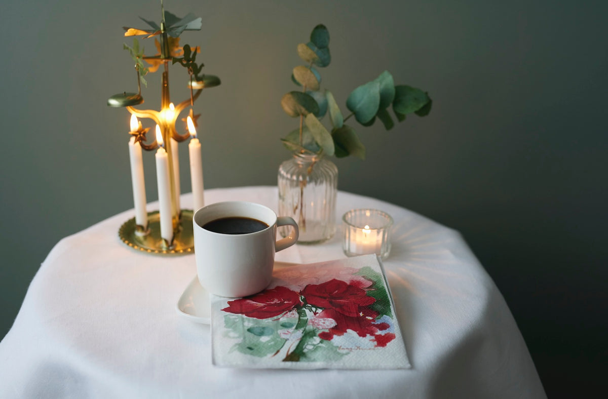 Gold angel chime with four lit Havi white candles, creating a warm glow on a white cloth against a dark green background.