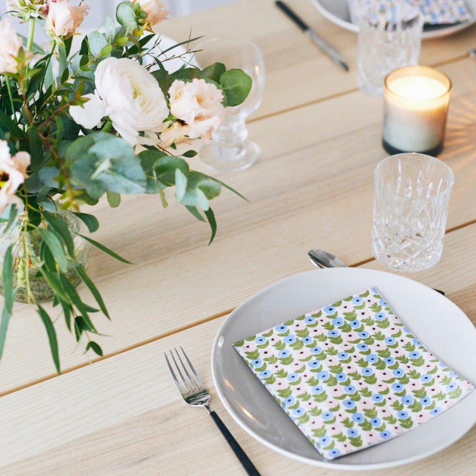 Havi Flora Napkin laid on a white dinner plate.   Clean wooden table is set for dinner with cutlery, glassware and and vase of flowers