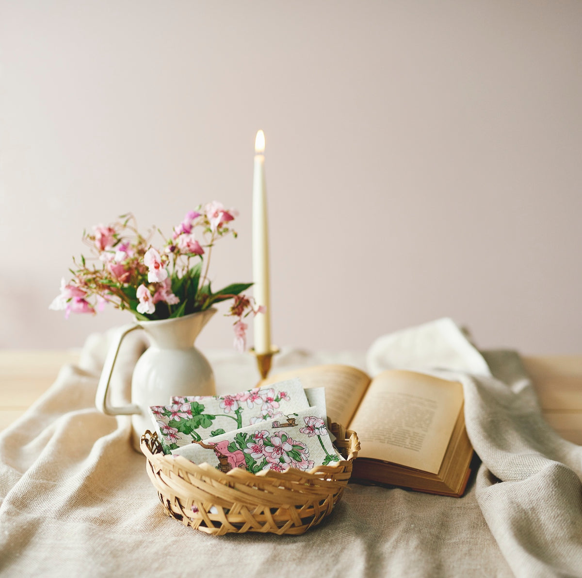 Havi Pelargonia paper napkins in a wicker basket, with a book and a lit candle and flowers