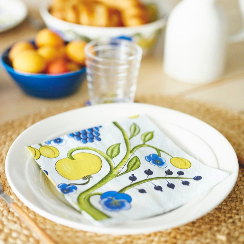 Tarha Paratiisi Havi Paper Napkin on a white plate, set on a dinner table with fruit and bread in the background