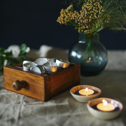 Havi Tuikku Tealights shown in a wooden box with two lit by the side.   Vase with greenery alongside. 