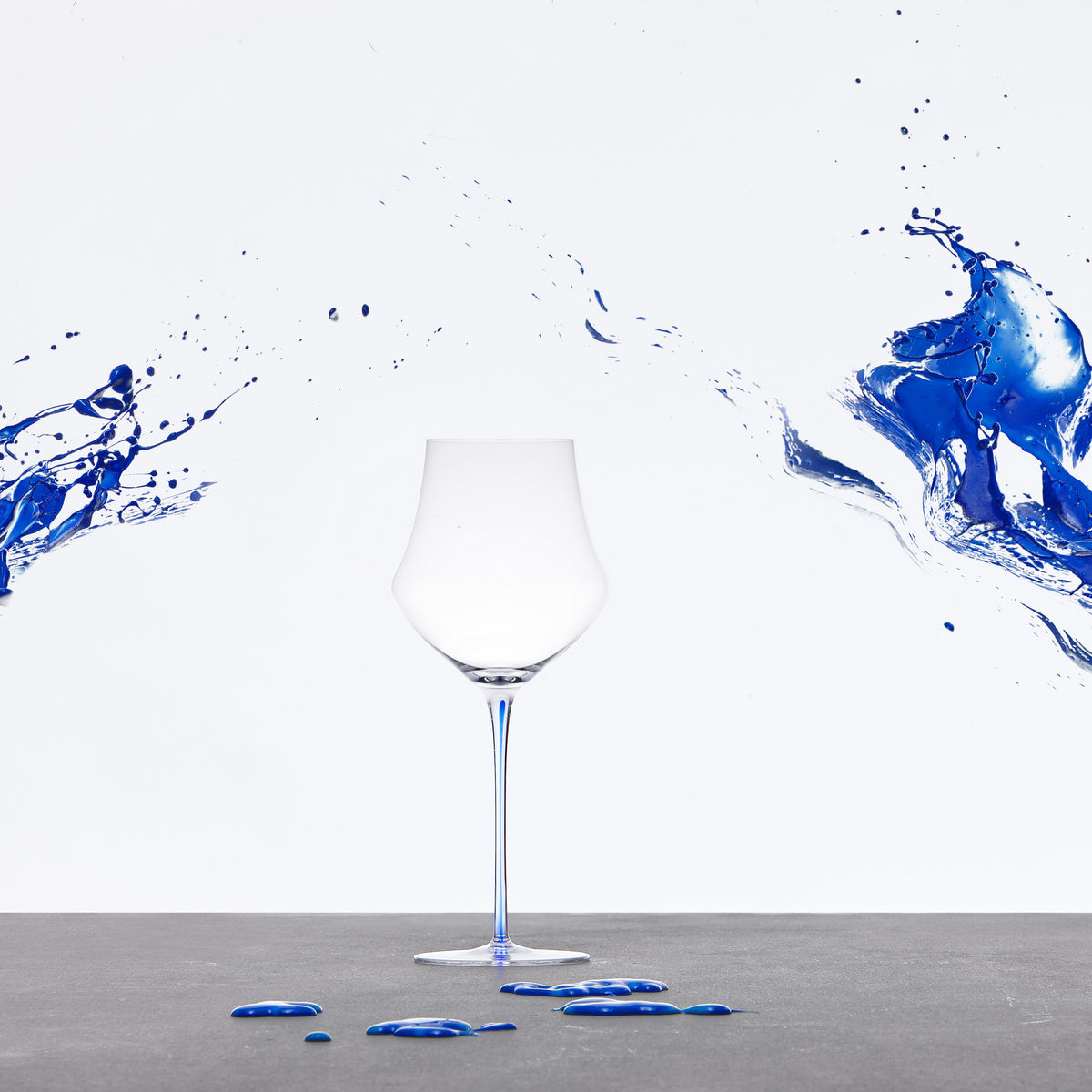 A clear Memento wine glass stands centered on a gray surface against a white background, with dramatic blue liquid splashes entering from both sides and small blue droplets scattered below.