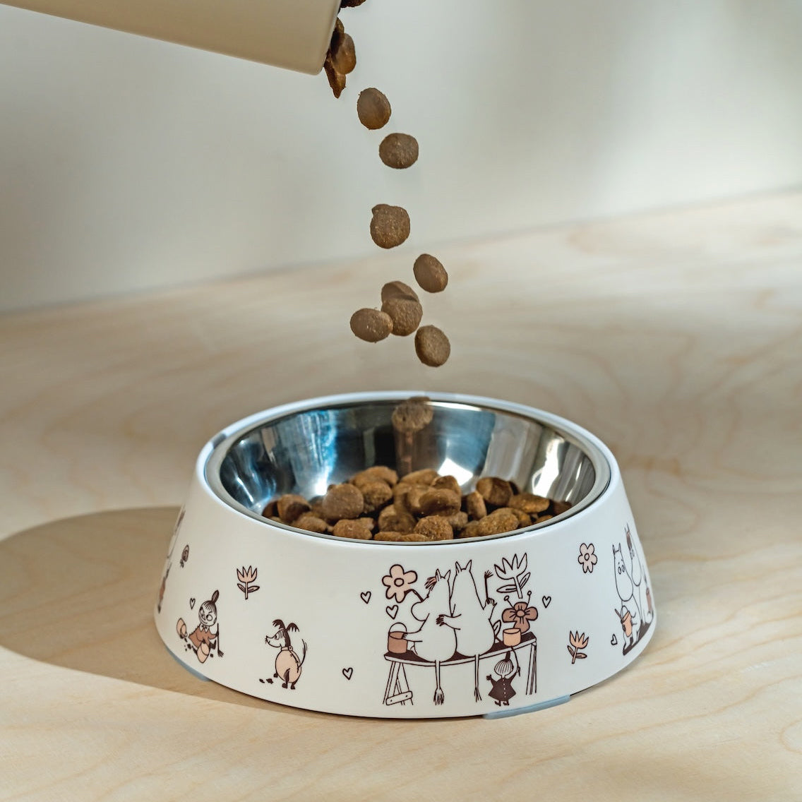 Dog food being poured into a decorative bowl with Moomin illustrations on a wooden surface.