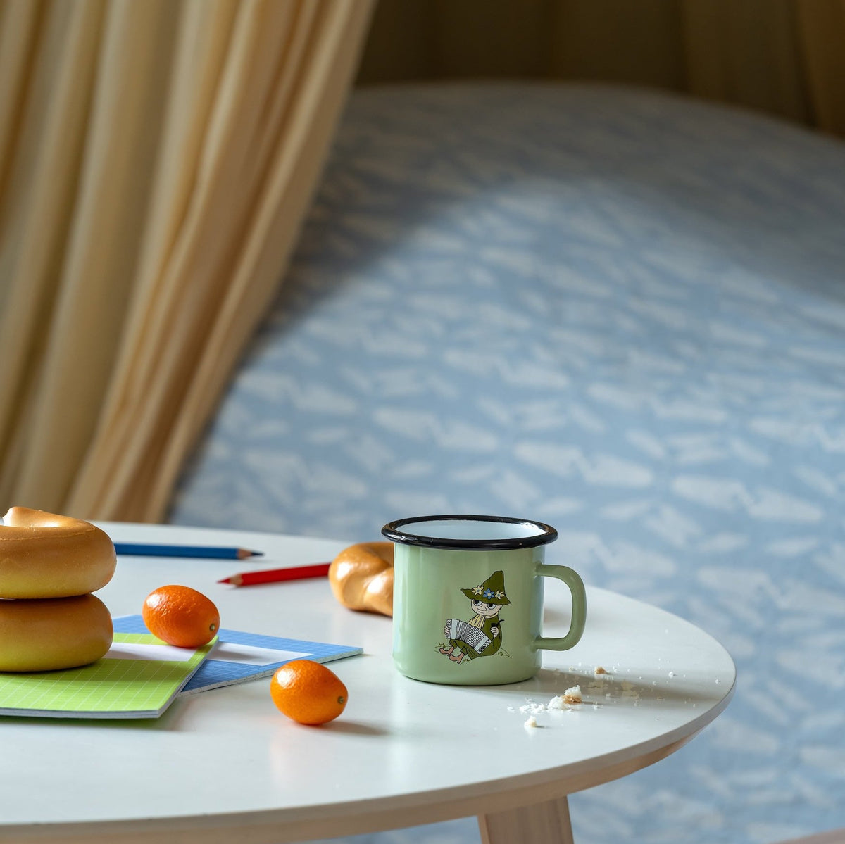 Muurla Moomin Snufkin Enamel Mug on a table next to oranges and breadcrumbs.