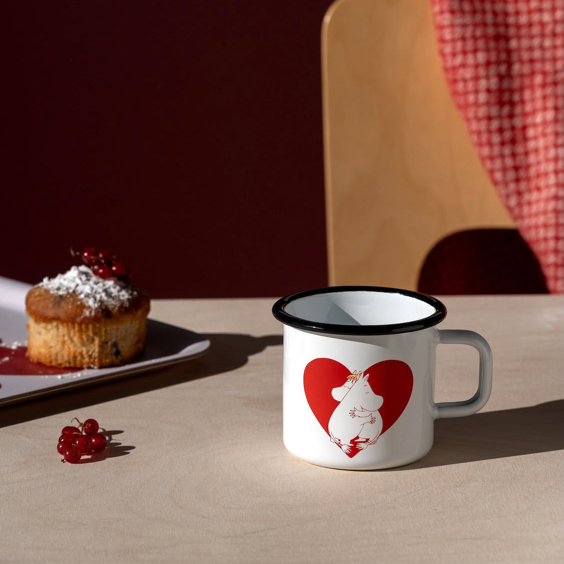 Mug with a heart design and Moomin characters on a table with a cake and red checkered cloth in the background.