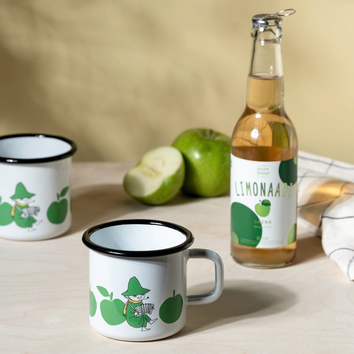 Two enamel mugs with green Moomin apple designs, a bottle of lemonade, and green apples on a light surface.