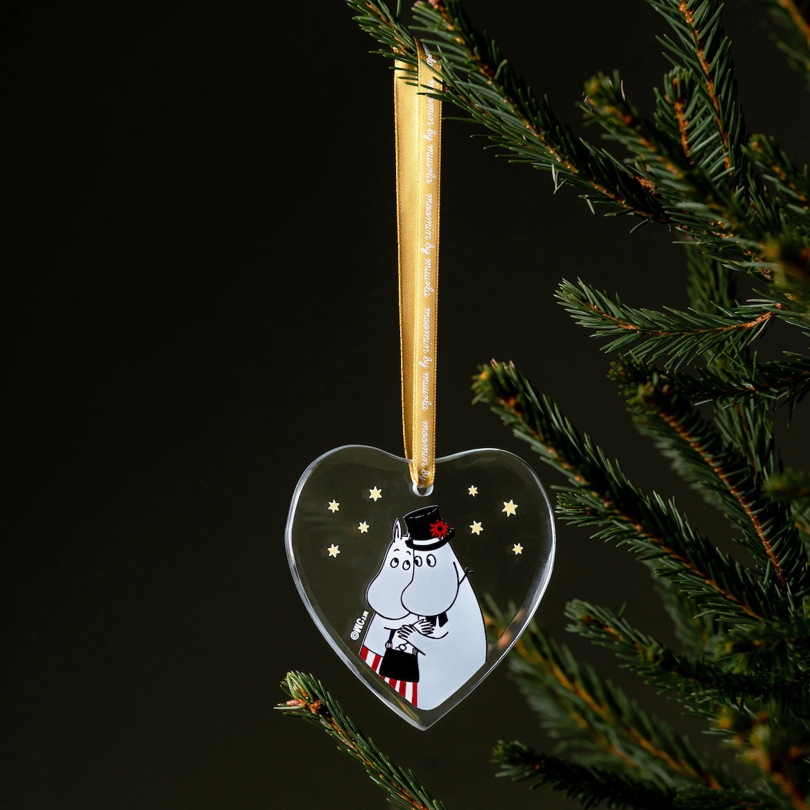 Heart-shaped Christmas ornament with a Moomin characters on a tree branch against a dark background