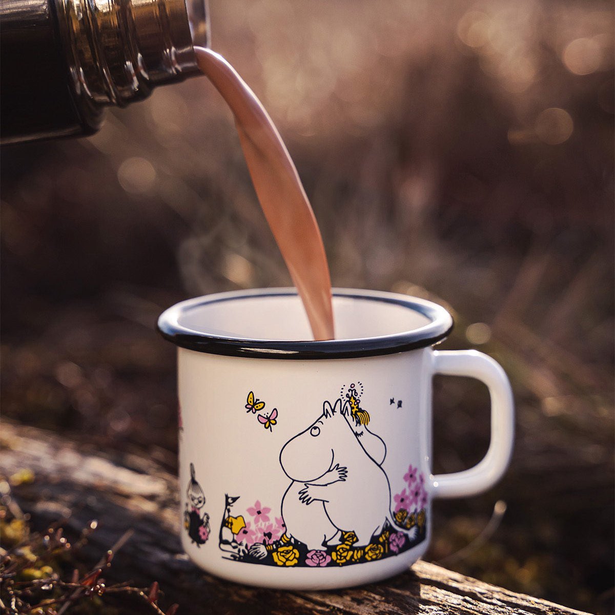 Coffee from a flask being poured into a Moomin Hug Enamel Mug, standing on a fallen tree.