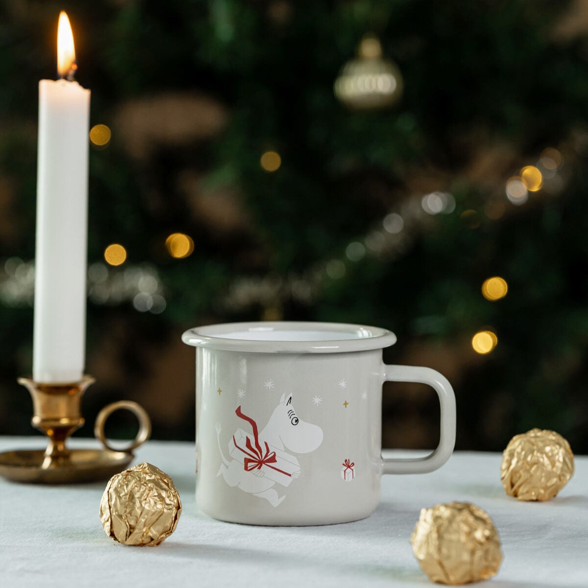 Moomin Gifts Enamel Mug 37cl, by Muurla Design, shown in a Christmas setting with candles , chocolates and a Christmas tree in the background