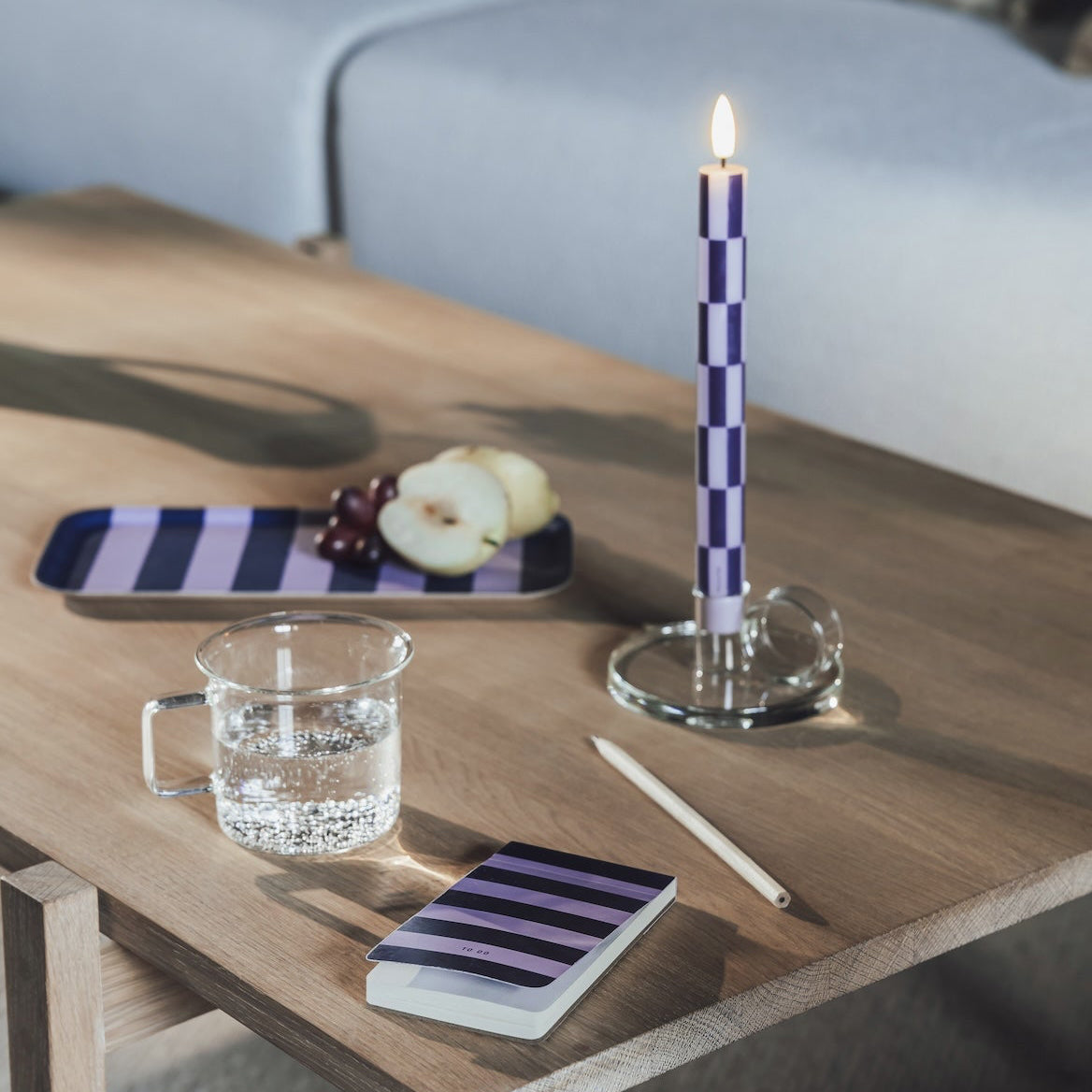 A lit purple and white checkered candle in a glass holder sits on a wooden coffee table in a modern living room setting, alongside a striped tray, a glass of water, and a notebook.
