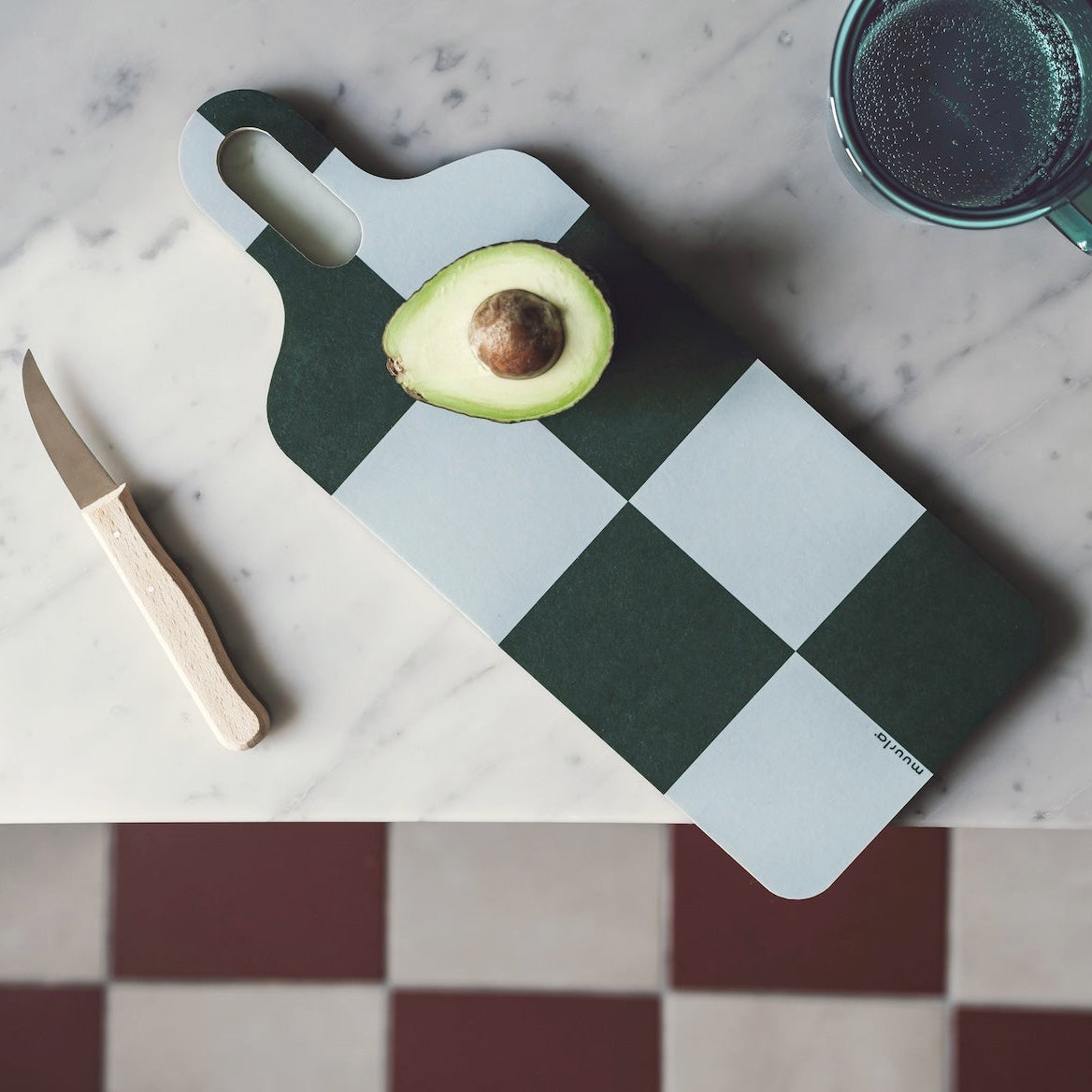 A dark green and white checkered cutting board with a handle, styled on a white marble countertop with a halved avocado, a small knife, and bok choy, viewed from above against a checkered floor.