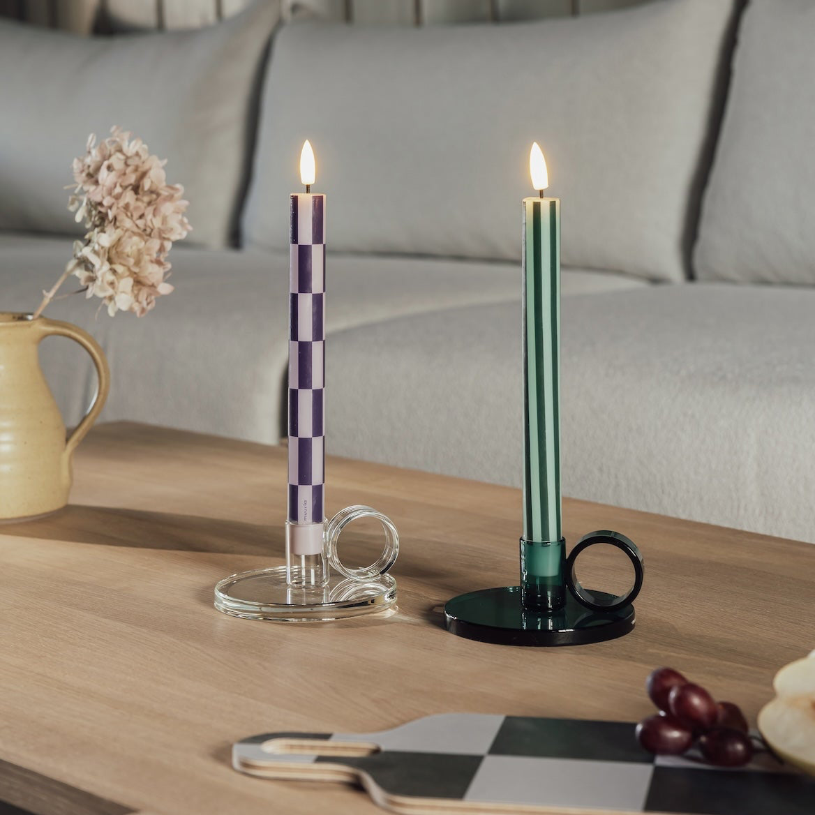 Two decorative Muurla taper LED candles in holders on a wooden table in a living room setting. One candle has a purple and white checkerboard pattern, and the other is dark green with a matching green holder.