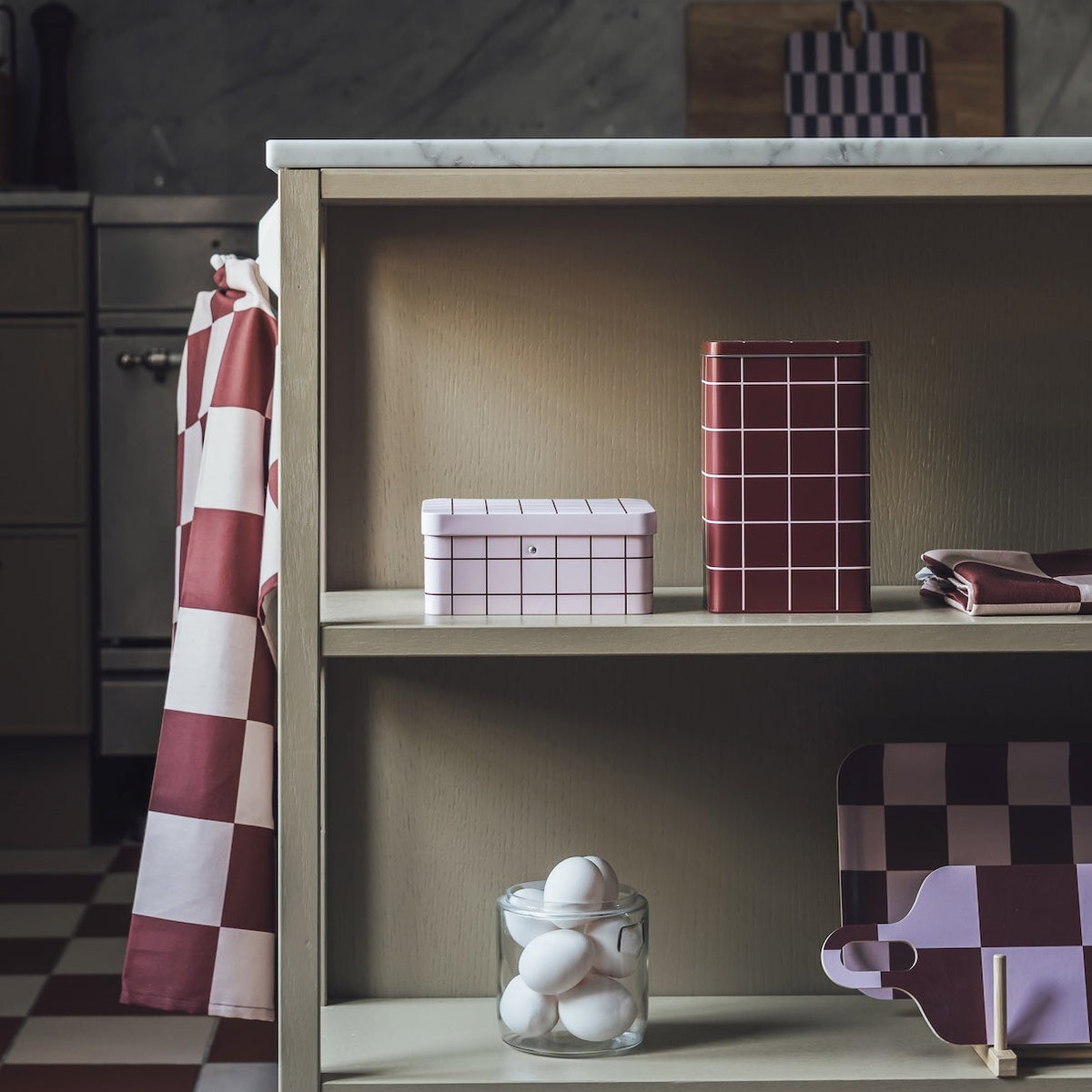 A rustic kitchen scene featuring a light wood open shelf unit with a white marble top, styled with patterned boxes and a small white ceramic figurine. A red and white checkered apron hangs from the side, set against dark wood cabinets and a checkered floor.