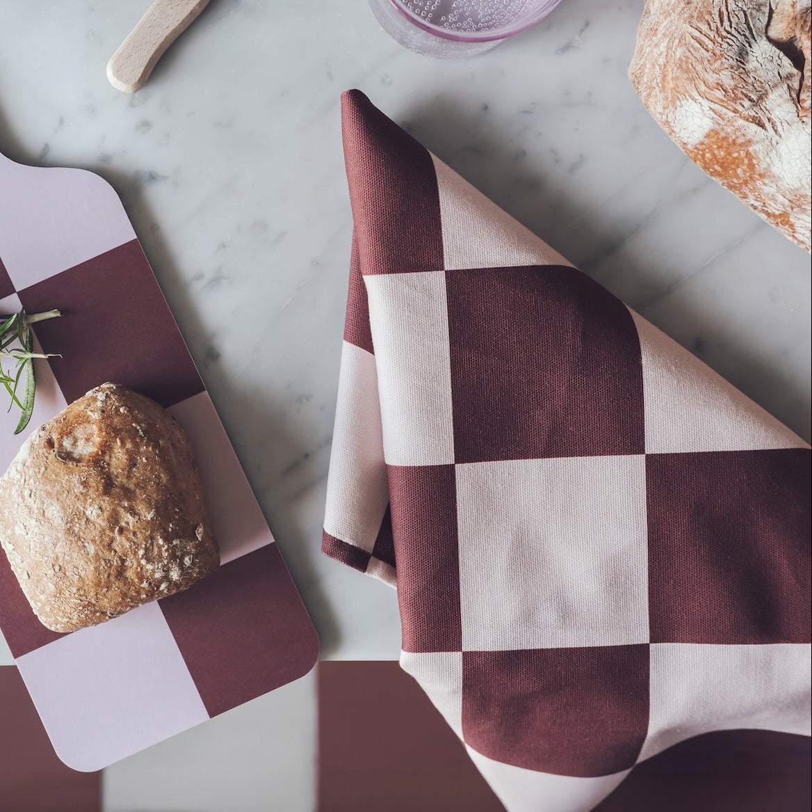 A styled flat-lay of a checkered kitchen cutting board and matching napkin in deep burgundy and cream, with a loaf of bread and a sprig of rosemary on the board, set on a white marble countertop.