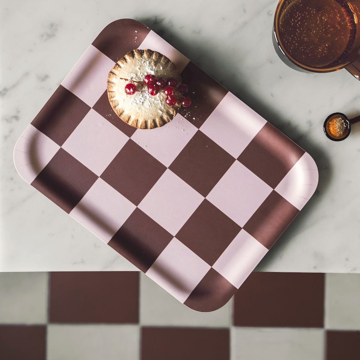 A rectangular Birch Muurla tray with a pink and dark brown checkerboard pattern, styled with a small pastry on top. The tray is placed on a white marble surface, with a checkered floor in the background and kitchen props like a mug, spoon, and spice jar nearby.