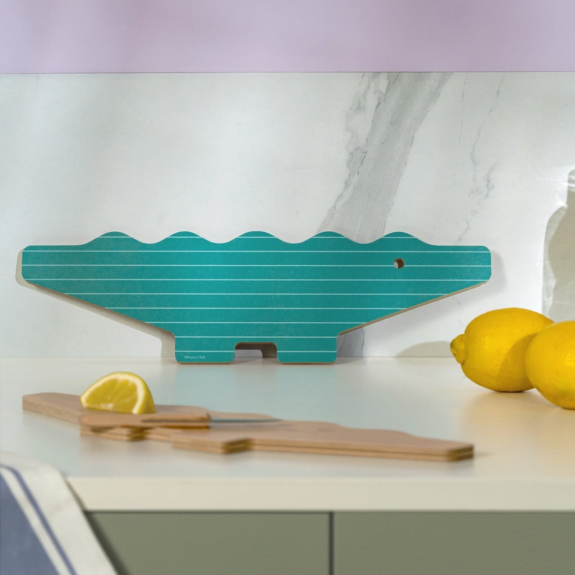 Teal alligator-shaped cutting board on a kitchen counter with lemons and a wooden spatula.