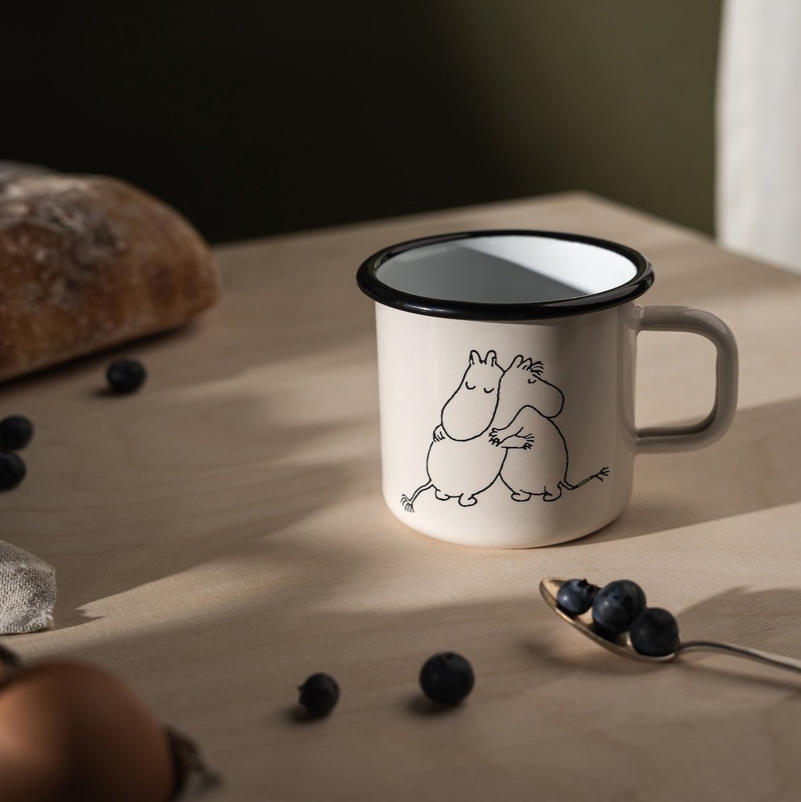 Moomin 80 Enamel Mug by Muurla Design.   Placed on a wooden worktop with bread, berries and a teaspoon next to it