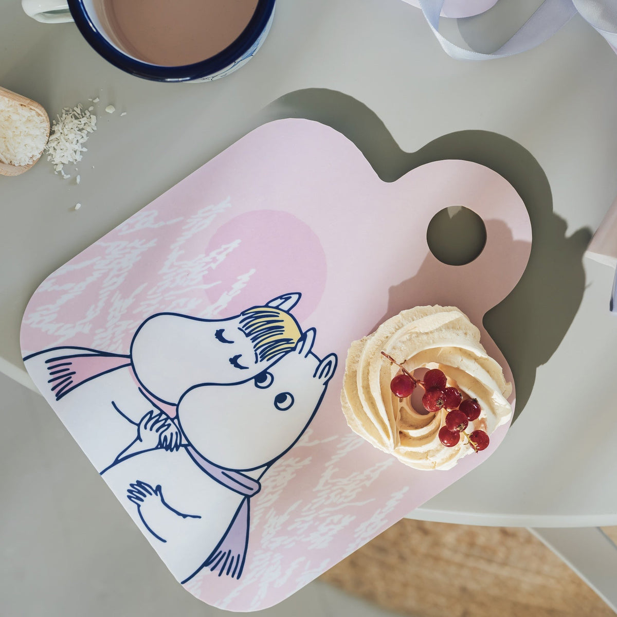 A heart-shaped pink Moomin cutting board with a Moomin character illustration, styled on a light gray table with a cup, sugar, and a pastry on top.
