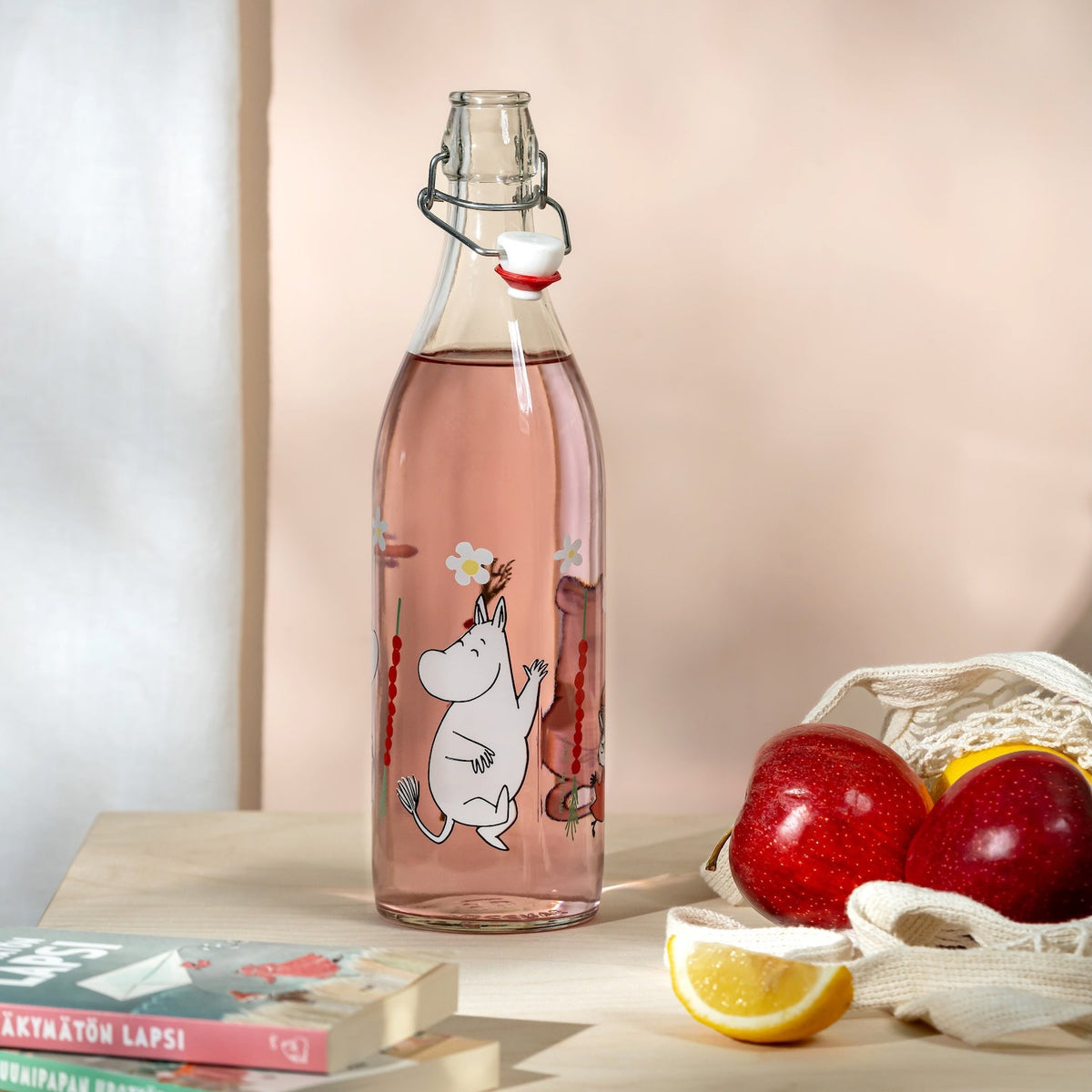 Glass bottle with a Moomin Wild Strawberries design on a table with books and fruits