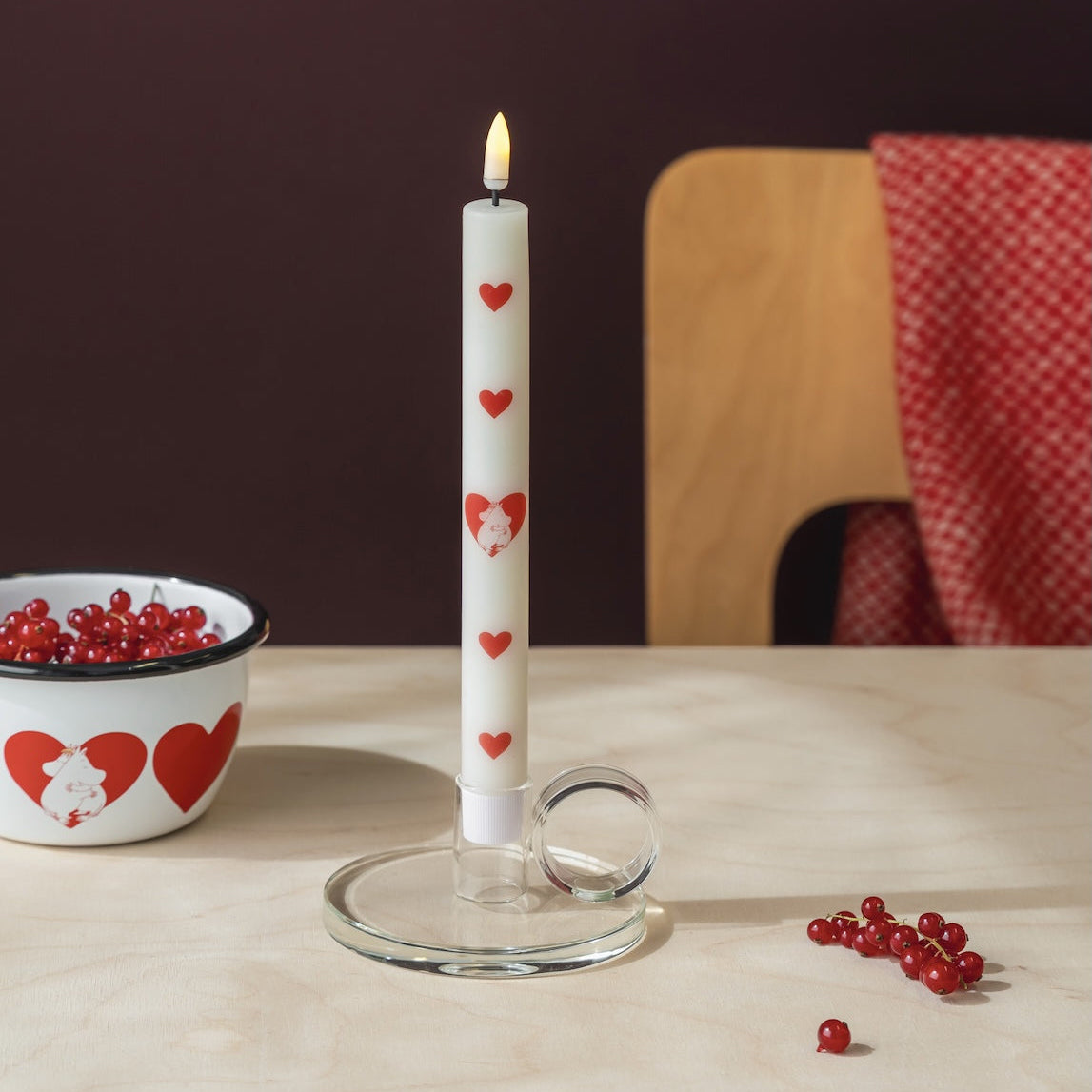 A white taper candle with red Moomin  heart motifs stands lit in a clear glass holder on a light wooden table. A matching white bowl with red hearts and red berries are visible in the background against a dark brown wall and wooden chair.