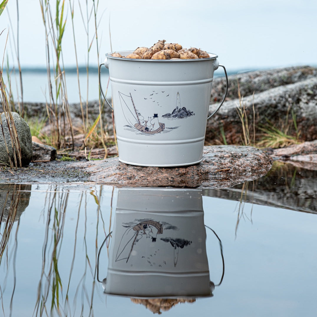 Moomin Sailors Bucket 10L shown next to the side of a pond, with a clear mirror reflection on the surface of the water 