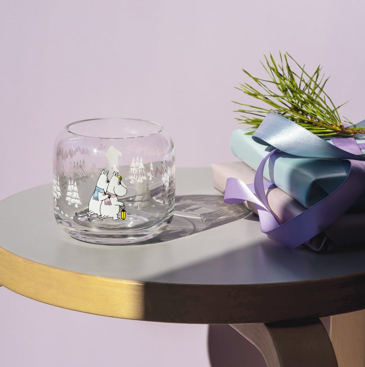 A clear glass tumbler with a Moomin winter forest illustration sits on a round wooden side table next to wrapped gift boxes tied with a purple ribbon and a small sprig of pine needles, against a soft lavender background.
