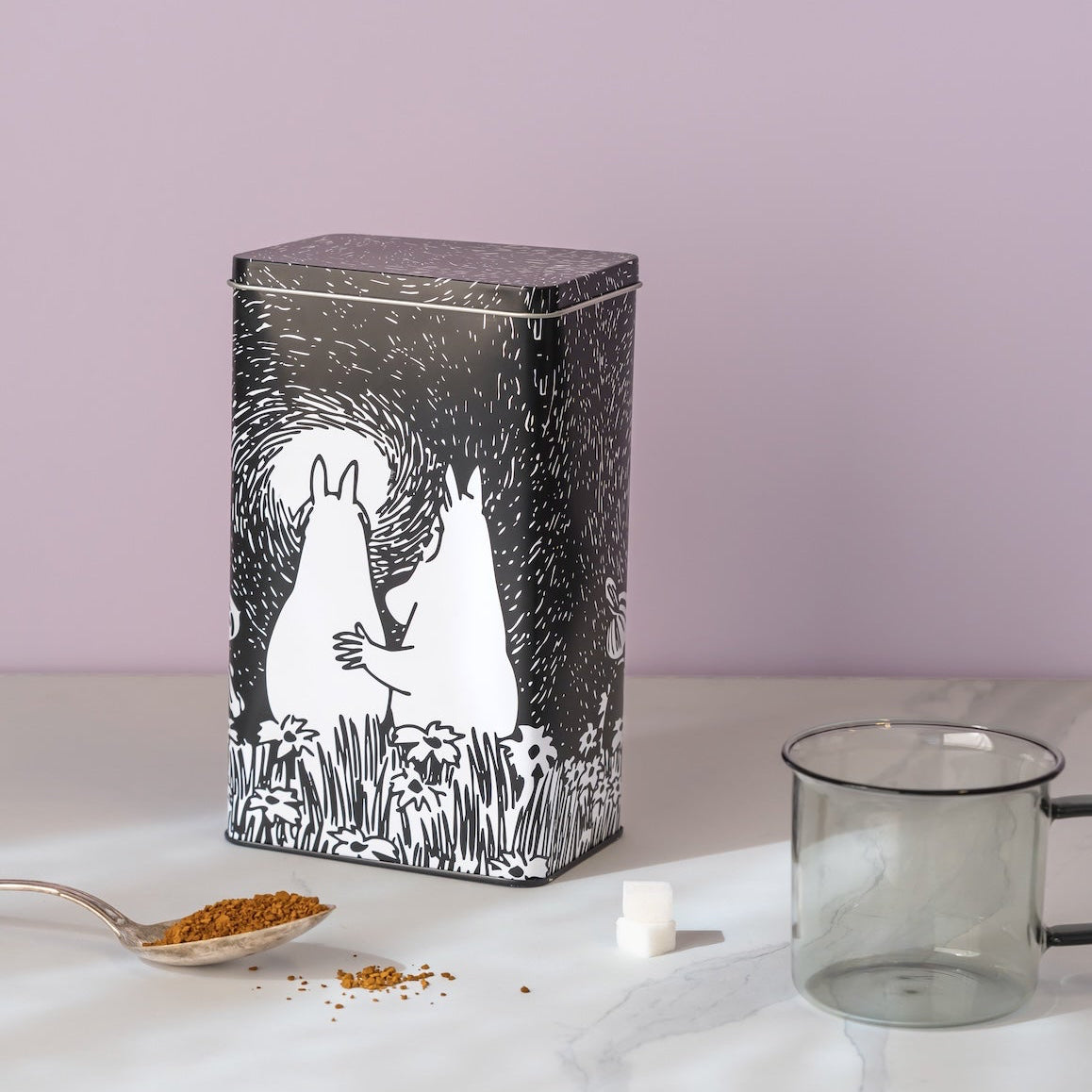 A rectangular Moomin coffee tin with a black and white illustration of two rabbits in a field, set on a white marble surface with a spoon of loose tea, a sugar cube, and a glass cup against a lavender background.