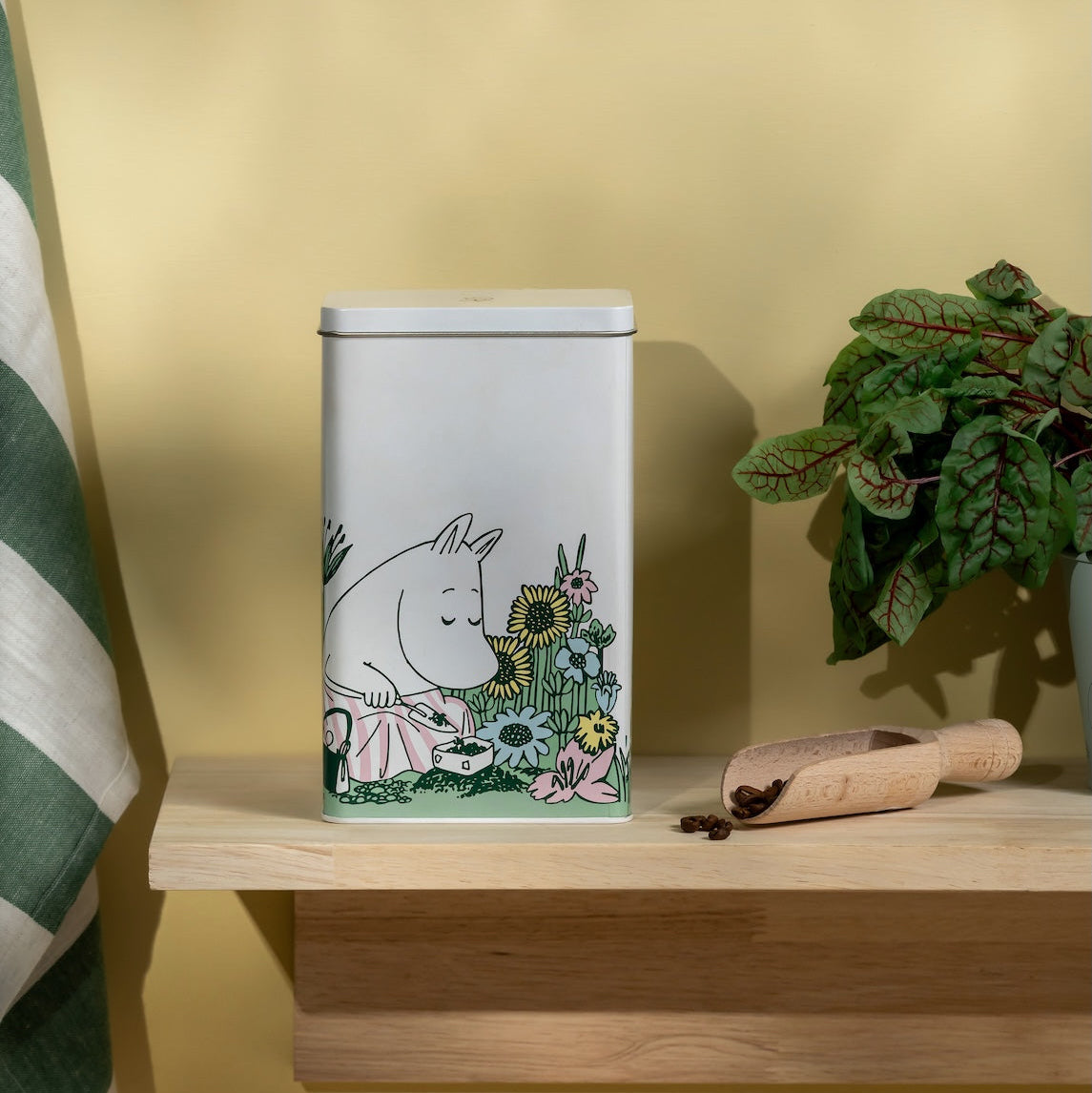 Moomin Decorative tin canister with a cartoon design on a wooden shelf against a yellow wall.