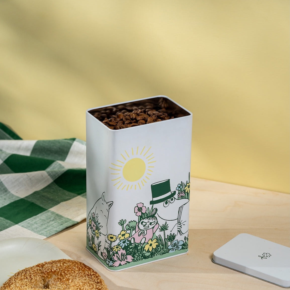 Moomin Decorative tin canister with a cartoon design on a wooden shelf against a yellow wall.