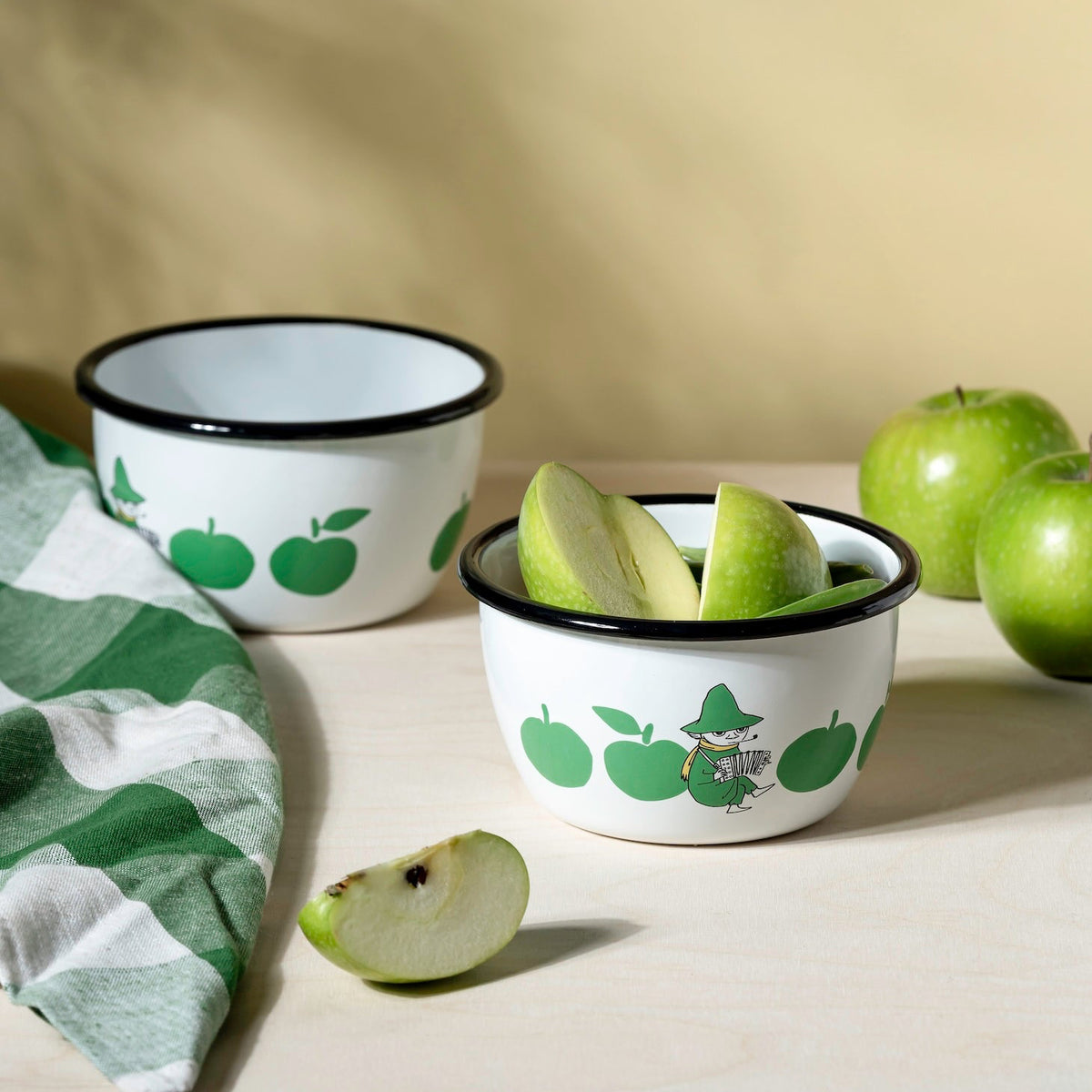 Two small bowls with Moomin green apple designs on a beige surface with green apples around.