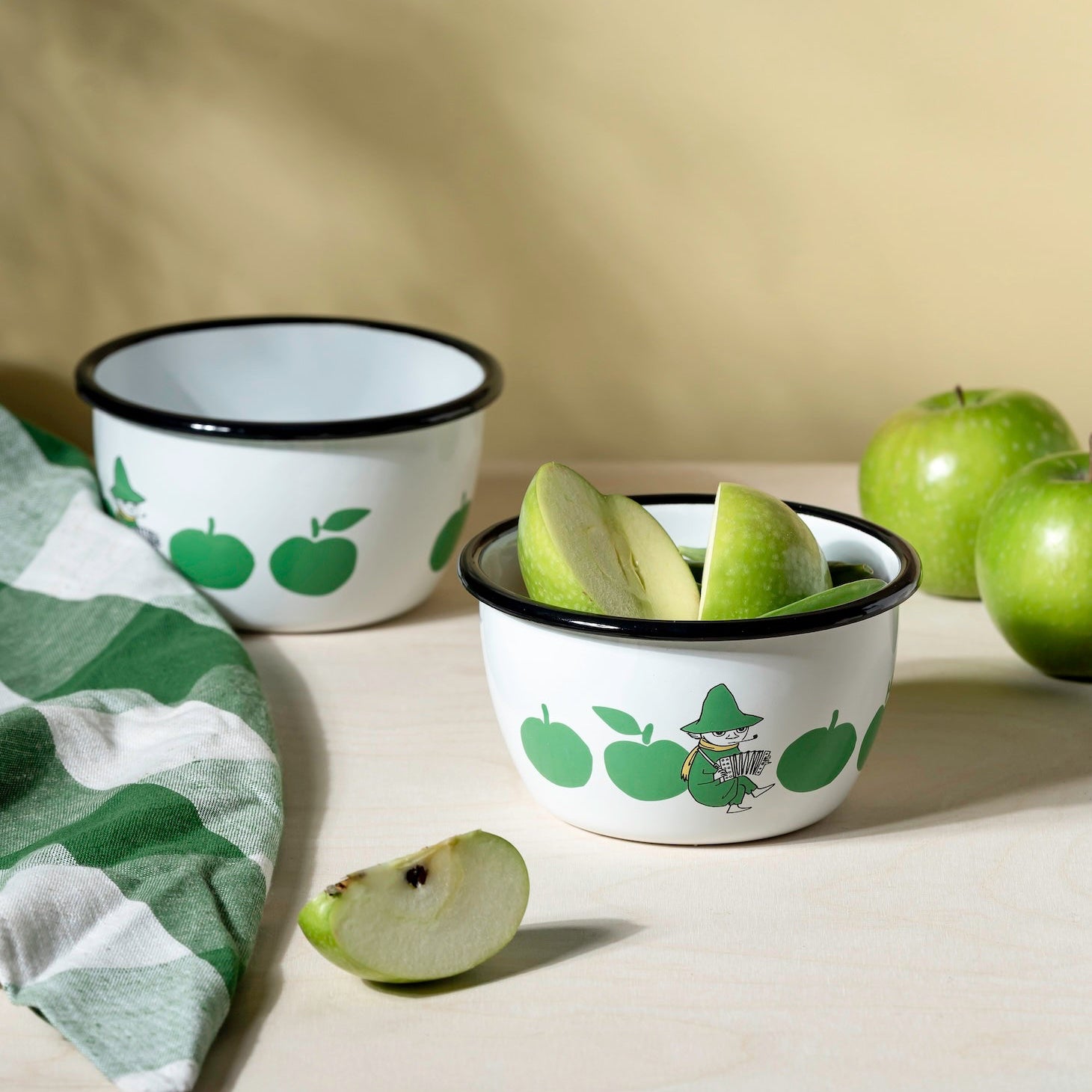 Two small bowls with Moomin green apple designs on a beige surface with green apples around.