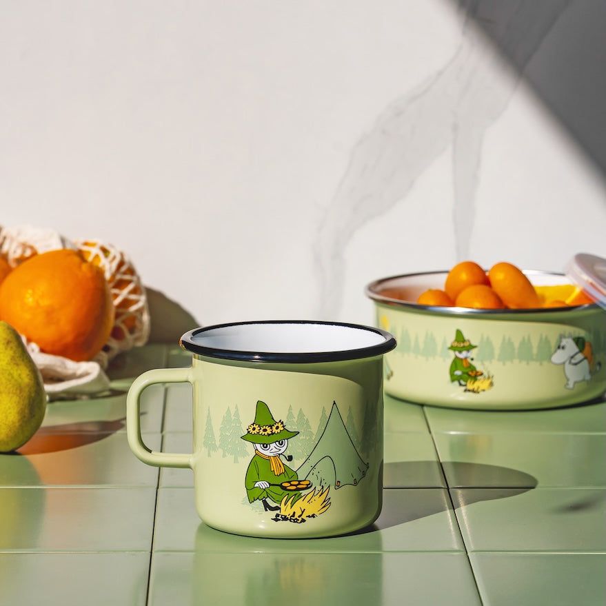 A light green Campers enamel Moomin mug with a cartoon camping character printed on it, set on a glossy green tiled countertop. In the background, another matching green bowl sits on a white surface with oranges and a pear nearby.