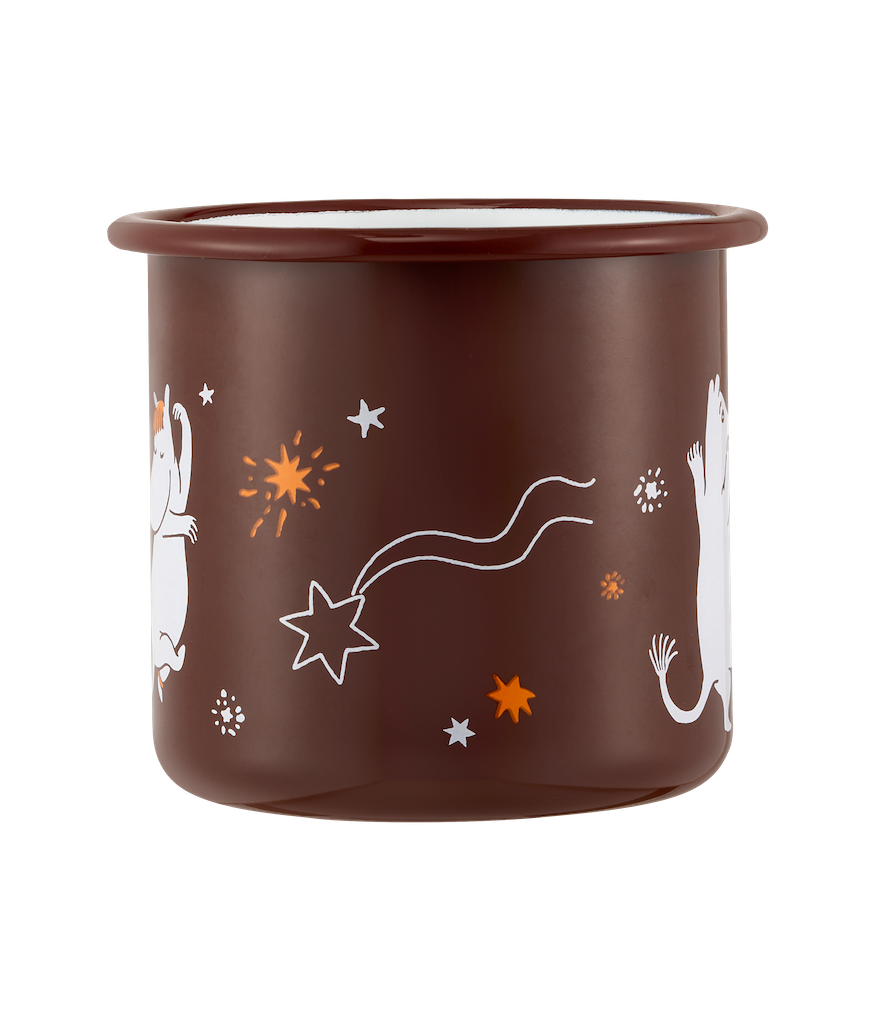 A dark brown Moomin Comet enamel mug with white Moomin character illustrations sits on a light marble table in a cozy indoor setting near a window, with a spoon of coffee and a plate of fruit nearby.