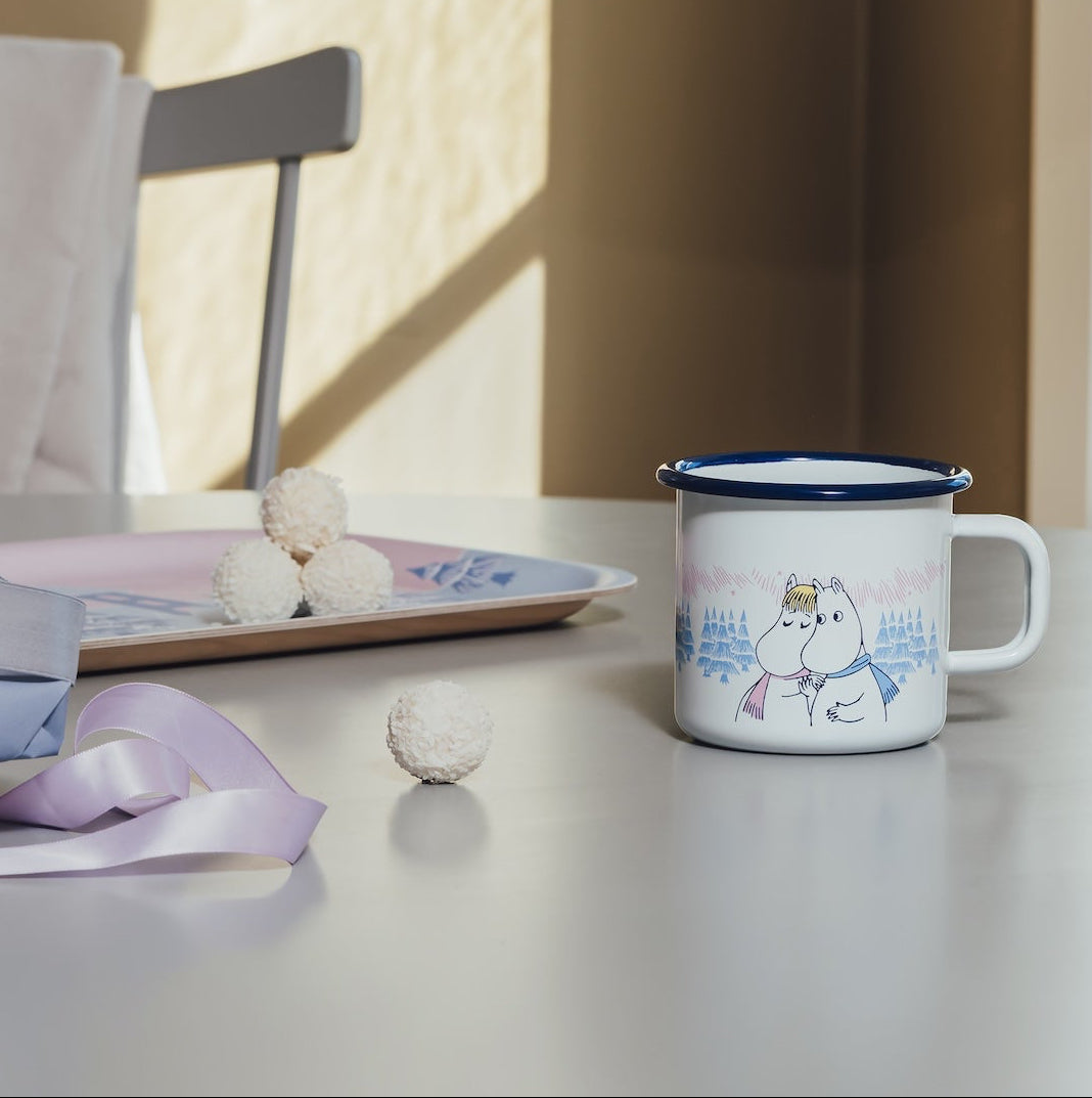 A white Moomin enamel mug with a blue rim and a winter-themed illustration of a snowman, styled on a tabletop next to a chair in a sunlit room.