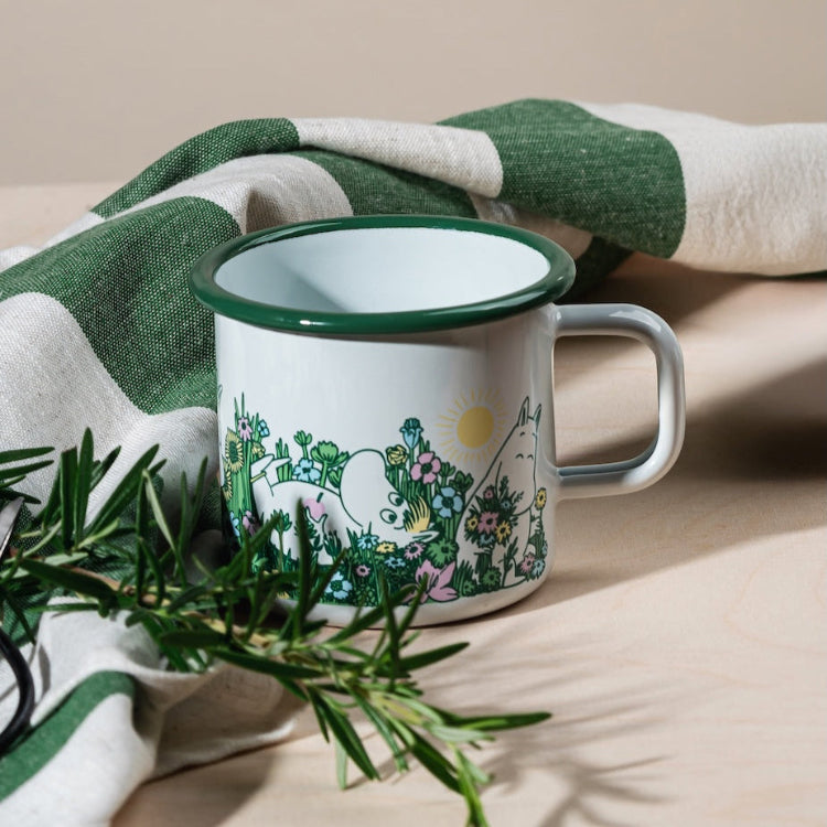 Moomin In the Garden Enamel Mug 0.37 L, pictured by a sprig of Rosemary and a green and white tea towel