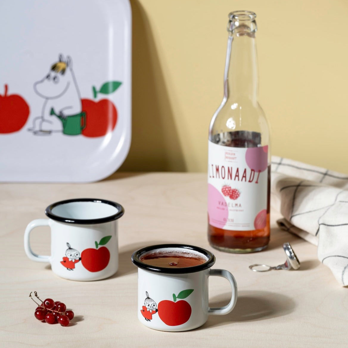 Two mugs with Moomin apple designs, a bottle of limonade, and a tray with a cartoon character on a light surface.
