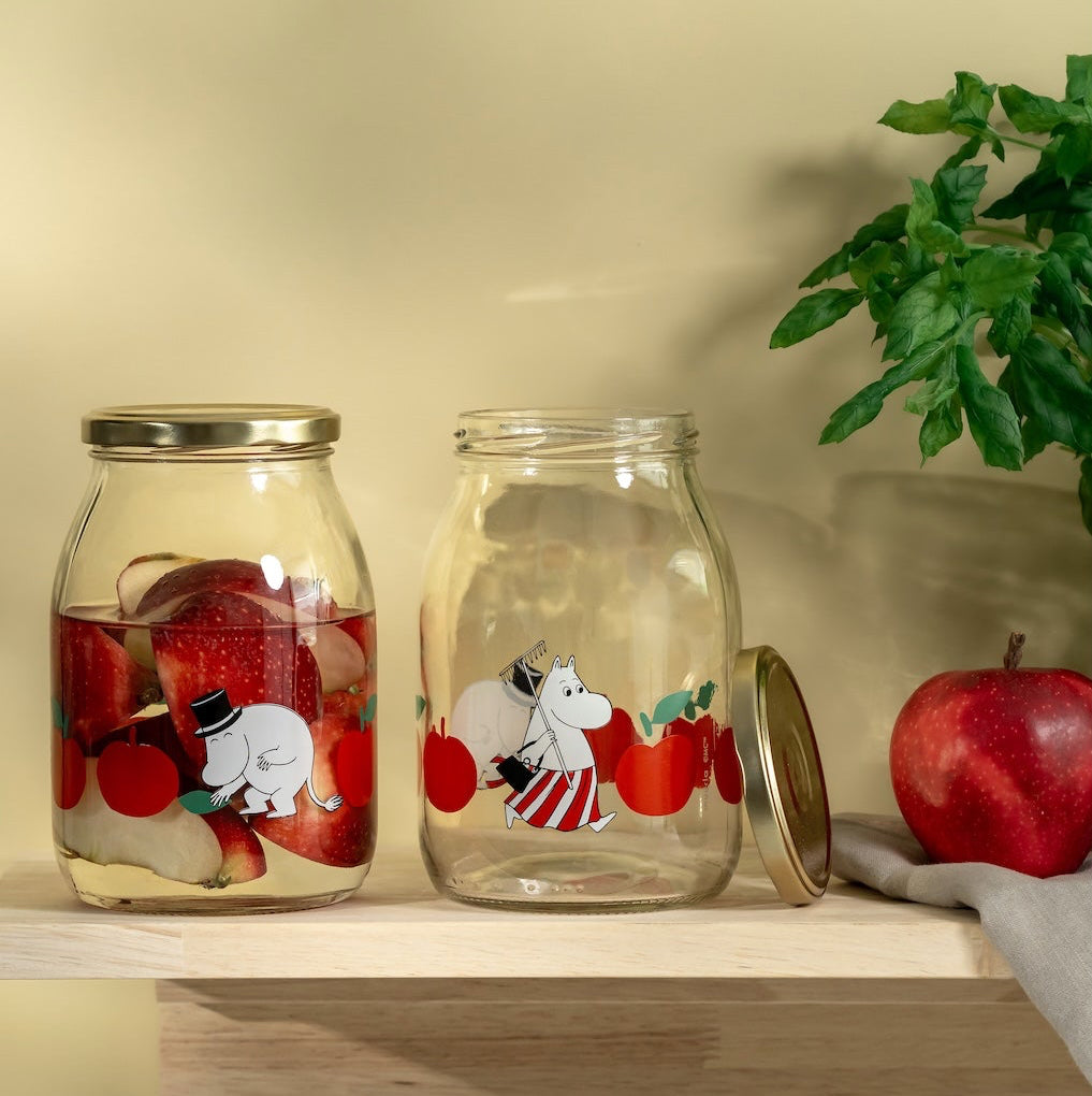 Two Moomin glass jars with illustrations of bears and apples on a wooden shelf against a beige wall.
