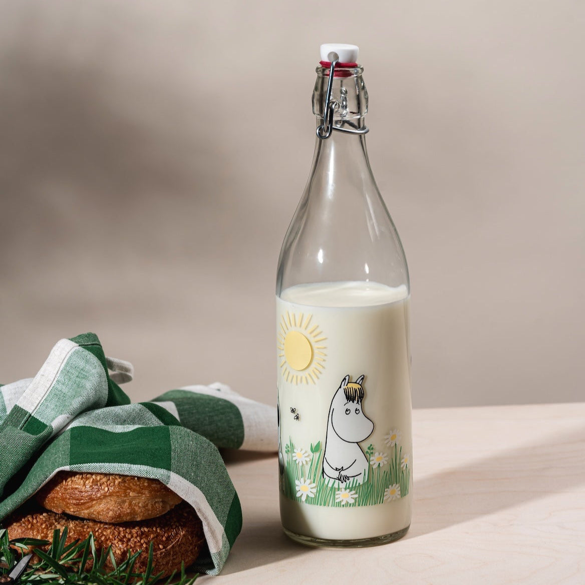 Moomin Meadow Glass Bottle with Clamp Stopper.  Pictured half full of milk, next to a green and white check tea-towel and some bagels!