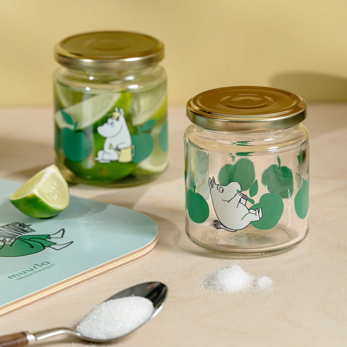 Two Moomin glass jars with illustrations on a beige surface with a spoon and lime wedge.