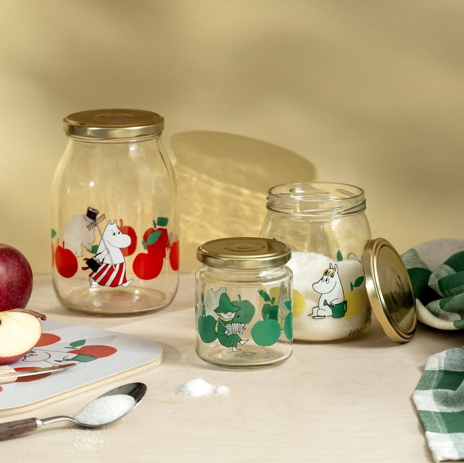 Glass jars with Moomin character designs on a table with an apple and spoon.
