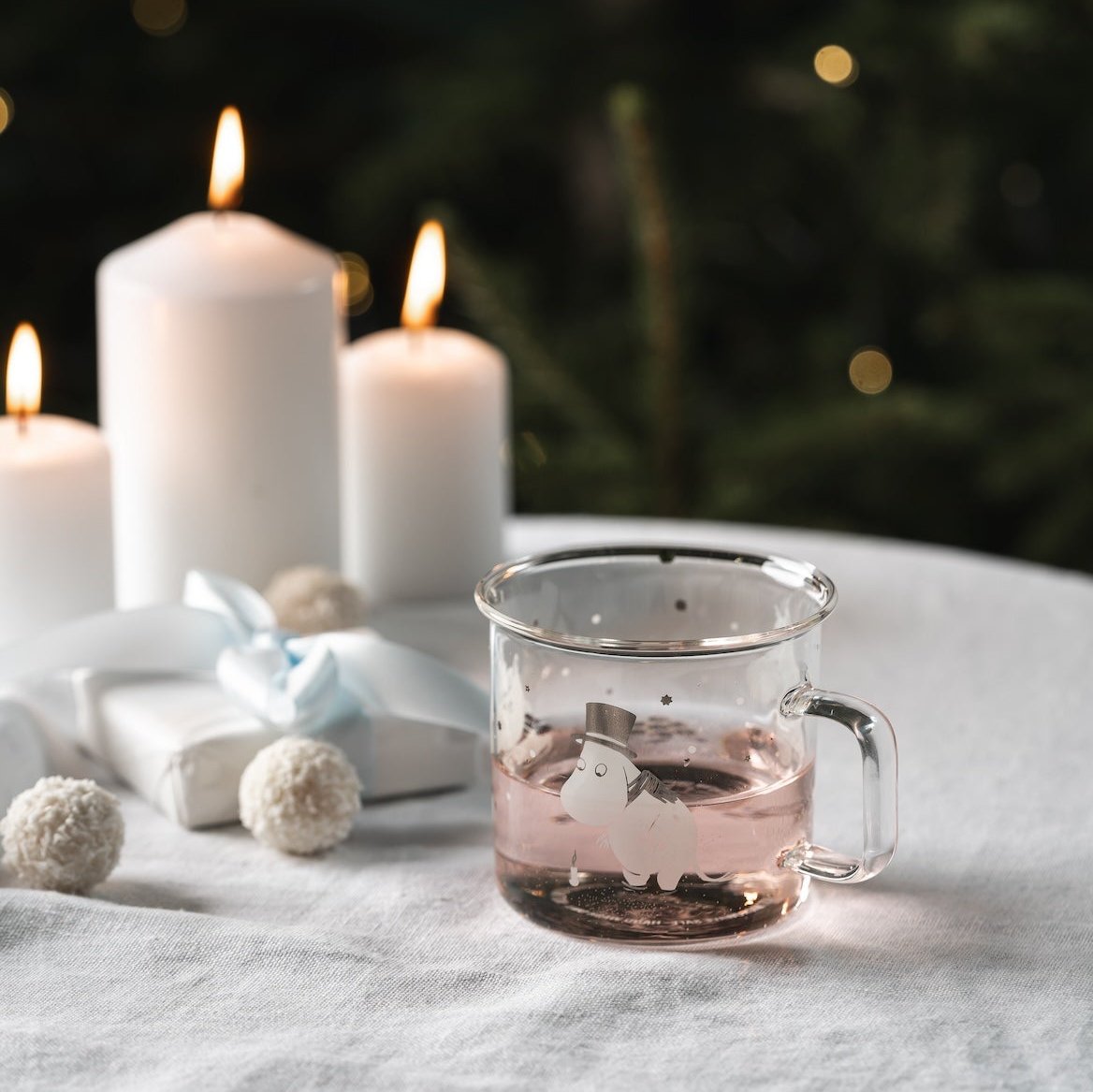 Moomin Let It Snow Glass Mug by Muurla, shown on a white tablecloth next to gifts and candles