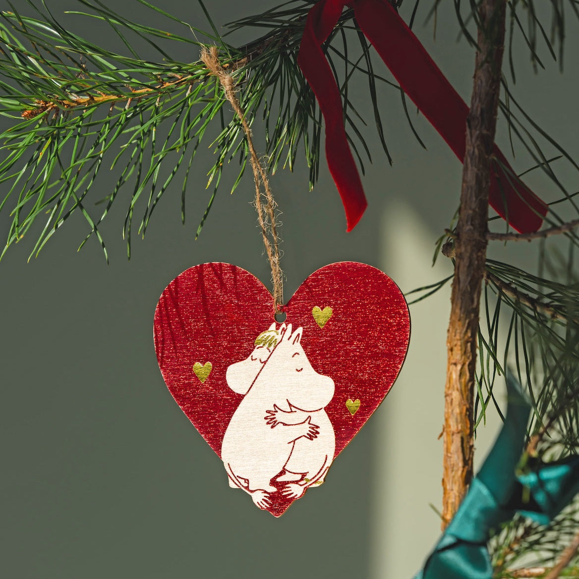 A heart-shaped Moomin wooden Christmas ornament featuring a white Moomin character, hanging from a pine tree branch with a red ribbon and other green pine needles in the background.