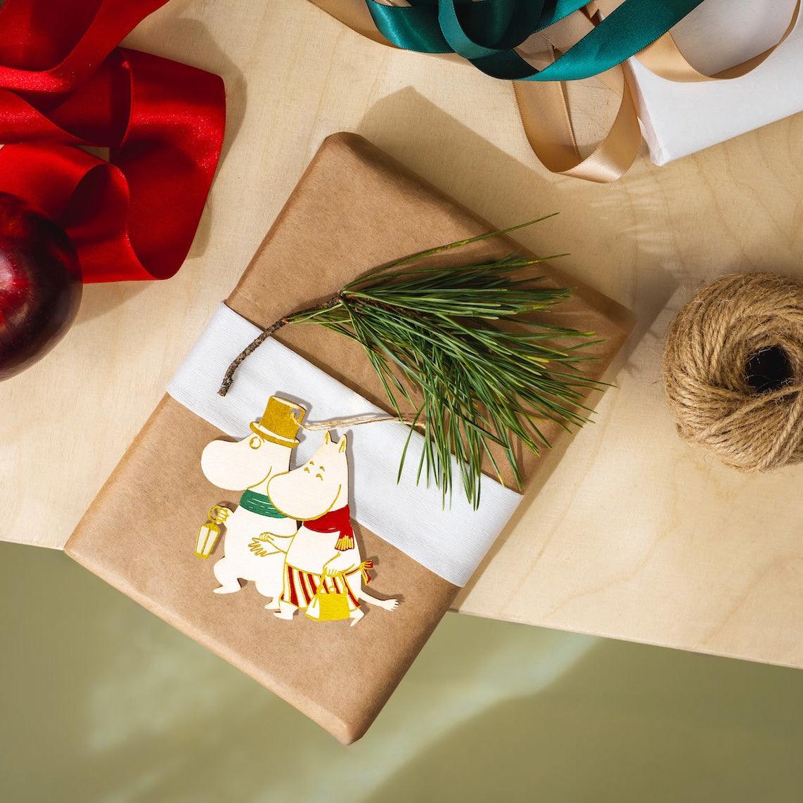 A styled flat-lay of wrapped gift boxes on kraft paper, with a red ribbon, a red apple, pine branches, twine, and a Moomin character ornament.