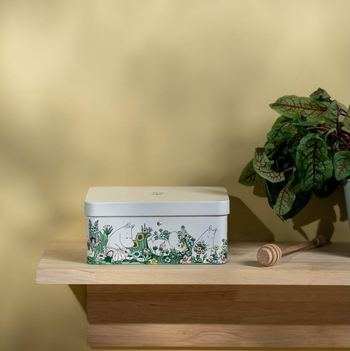 Moomin Decorative tin box with cartoon characters on a wooden shelf against a yellow wall.